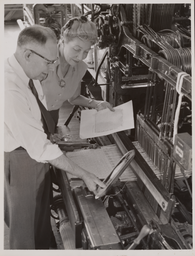Black and white photograph of a man and woman standing in front of an industrial loom; the man holds a shuttle and the woman holds a sketch.