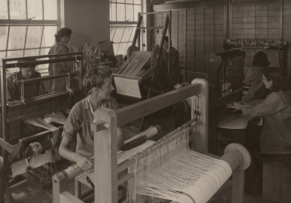 black and white image of a room filled with several looms. in the foreground Dorothy Liebes is seated at the loom.