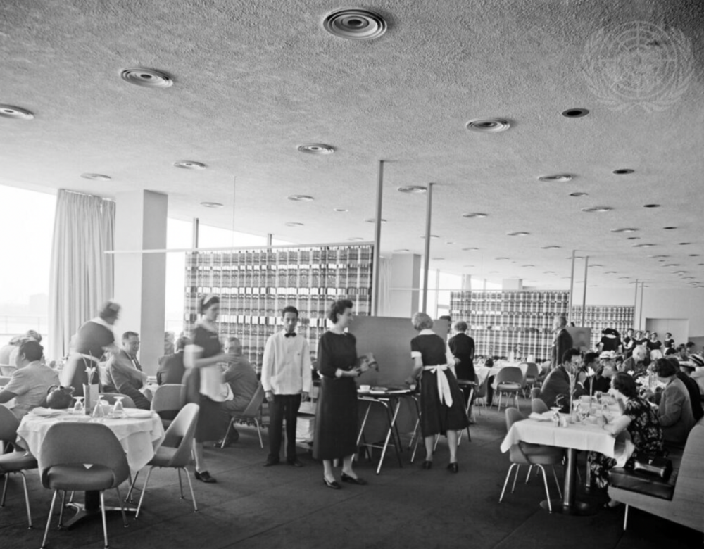 View of a large restaurant dining room with views of the East River through the floor-to-ceiling windows; handwoven screens are fully extended to create semiprivate dining areas in the large, open-plan space.