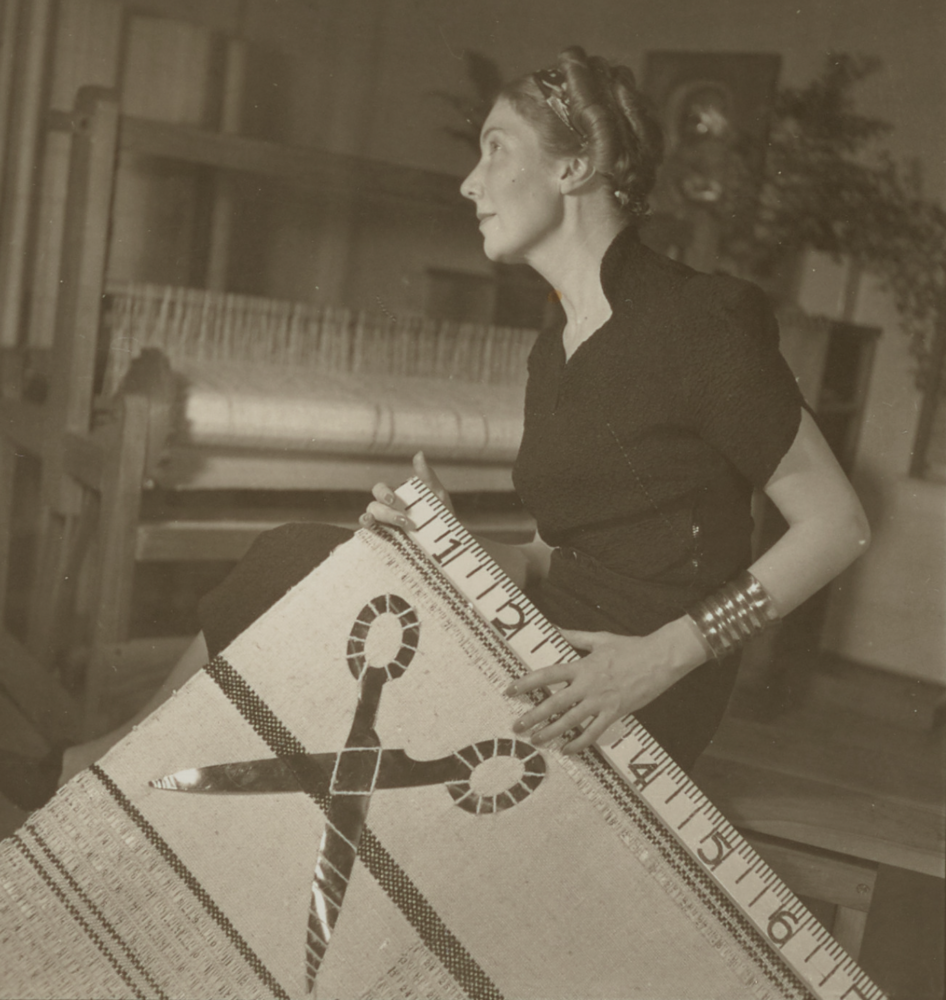 black and white photograph of a woman holding a large drapery. the drapery has metal scissors on it and a yardstick along the top