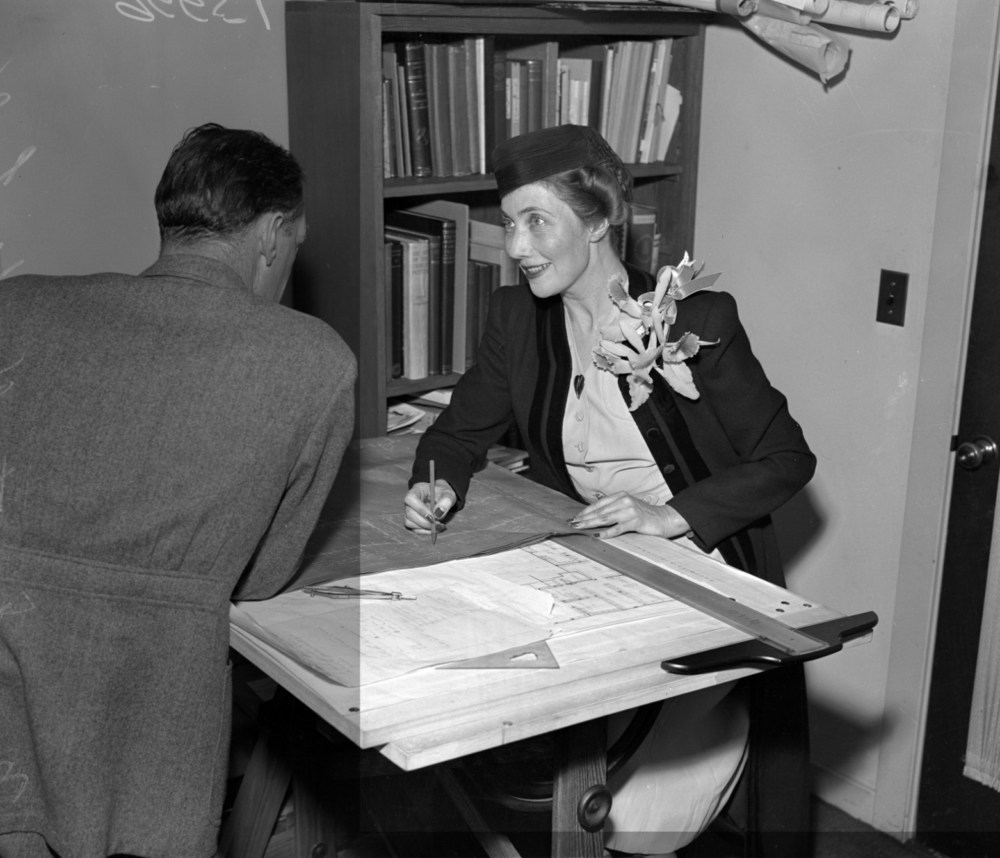 black and white photograph of a man and woman. the woman is seated and wearing a hat and her lapel has a large brooche on it. she has blueprints on the table and is poised with a pencil in hand.