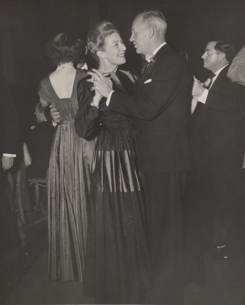 black and white photograph of a man and woman dressed in formalwear. they are holding hands engaged in a dance and are gazing lovingly at each other.