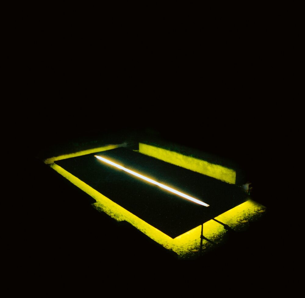 Three-quarter view of a black rectangle with a line of white light running along the middle; yellow light glows from beneath the rectangle. The rest of the image is all black.