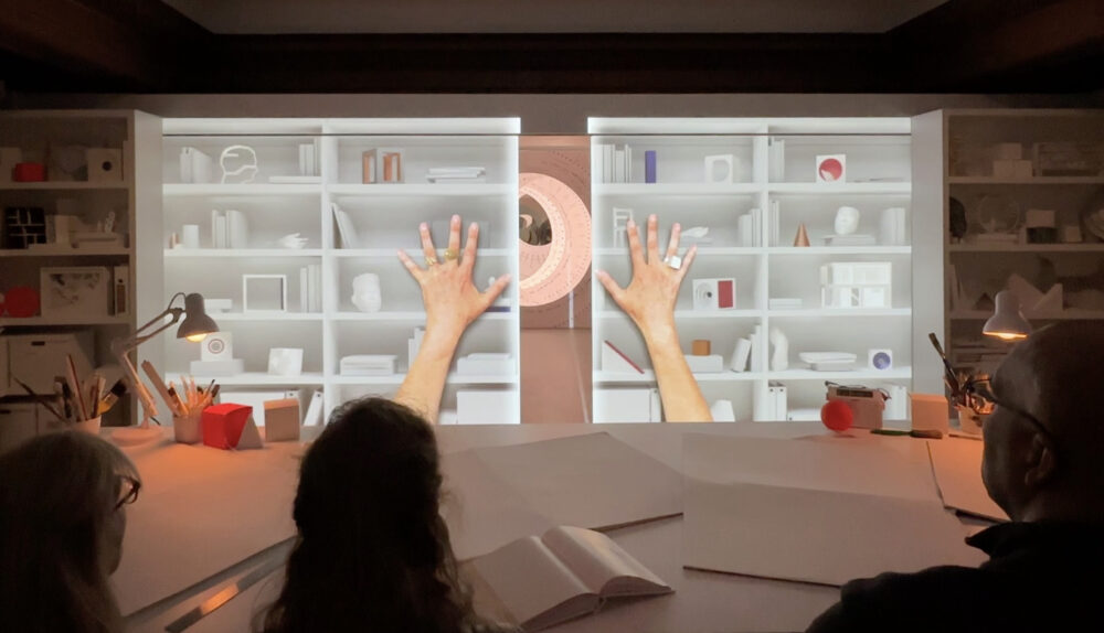 Three people sit before a table covered in open books, cups of pencils, and lamps. The people face a wall on which is projected imagery of shelves covered in a variety of objects overlaid by two outstretched human arms.