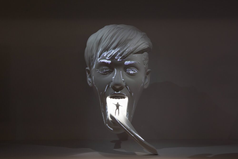 White model of a face with short hair and mouth wide open facing the viewer; a long tongue extends from the base of the mouth downwards to the right. White light is glowing from inside the model revealing a small figure standing at the start of the tongue.