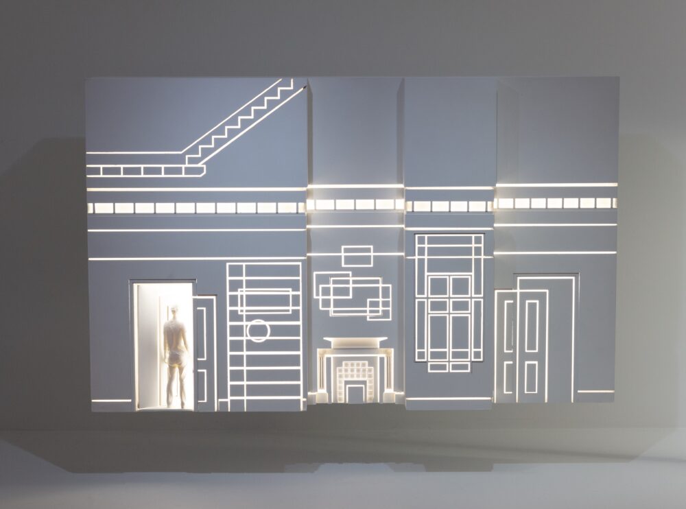 White rectangular model decorated with architectural forms which are revealed by the white light glowing from inside; a figure enters the model through a rectangular opening in the bottom left corner.