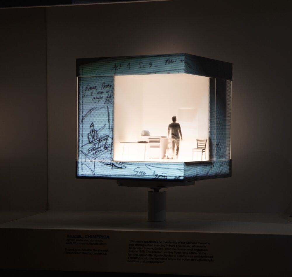 Installation view of a cubic model with an illuminated white interior space; projections of drawings illuminate the exterior. A figure stands in the interior space.