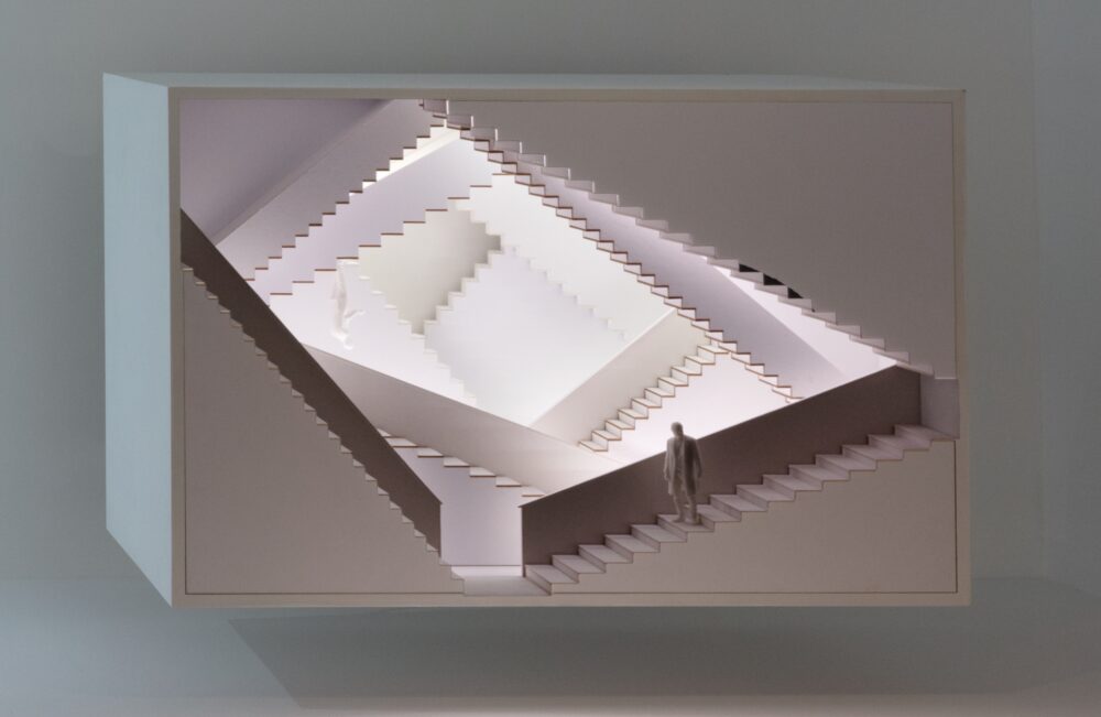 White rectangular model made of repeating layered staircases at different angles; two small figures are standing on different staircases.