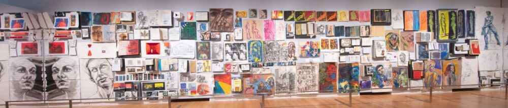 A wall of a gallery is covered with drawings and paintings of various sizes and colors.