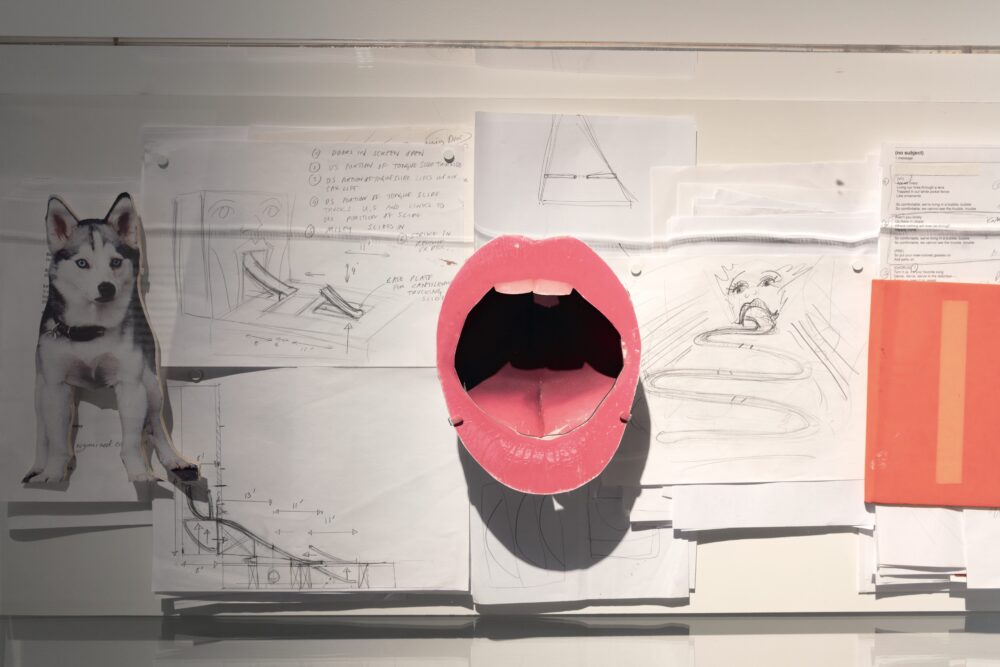 Paper cutout of an open mouth with pink lips foregrounds a collage of drawings pinned to a wall.