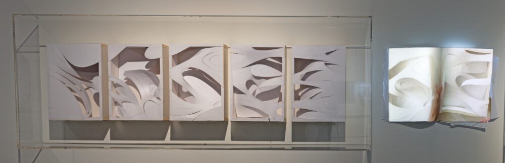 A clear case housing five white, rectangular sculptures with abstract cutouts is attached to a wall. A projection of a similar form as the cutouts glows to the right of it.