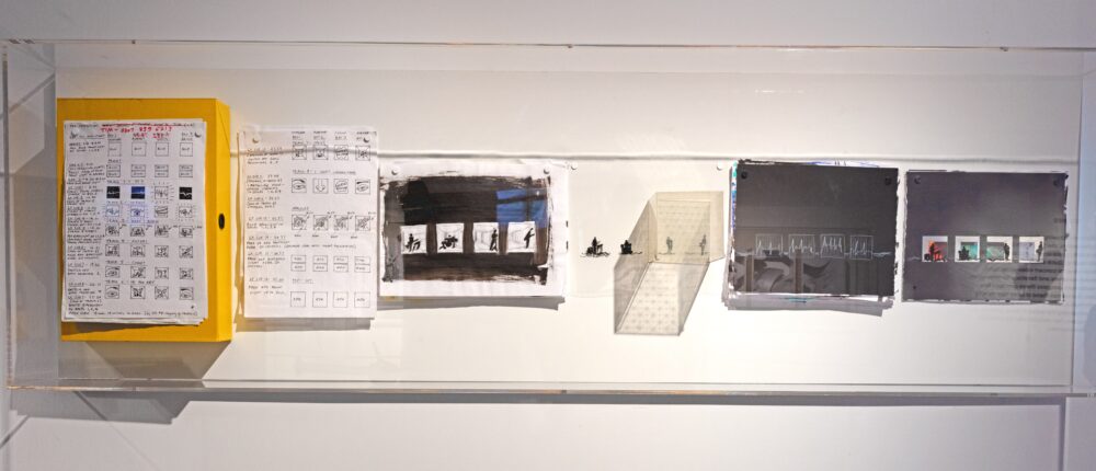 Six black-and-white drawings and a clear cube arranged horizontally within a display case