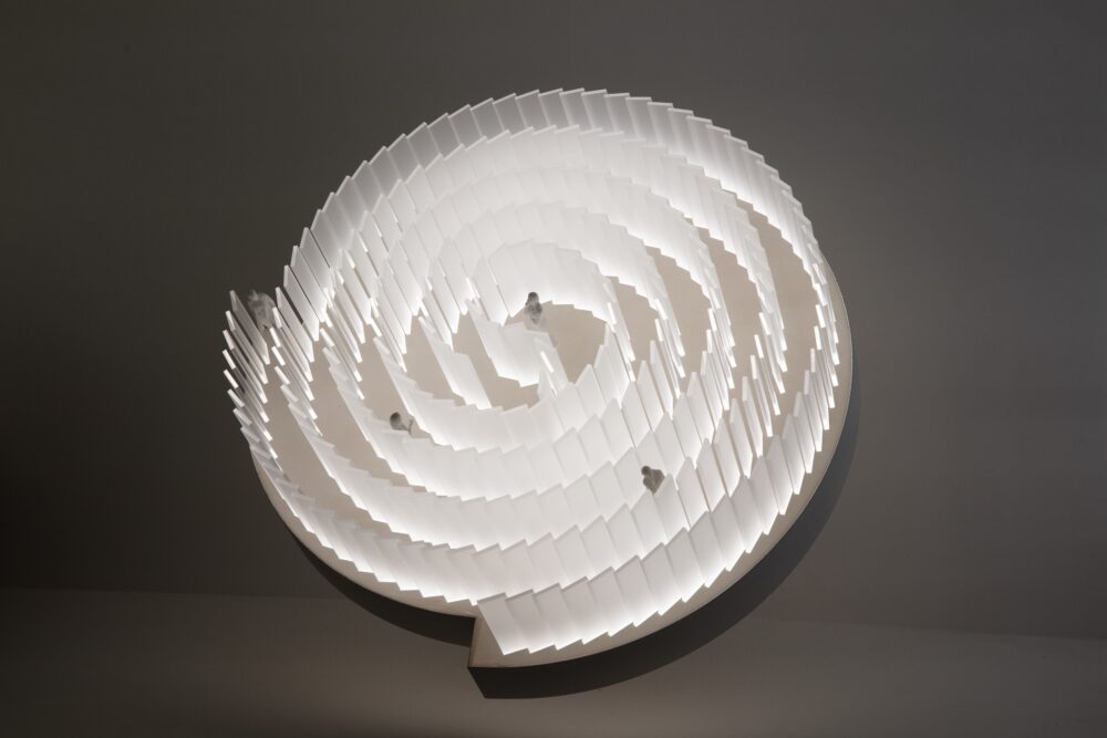 White model of spiraling pathway divided by curved translucent panels; a white light glows from within the model illuminating three figures walking along the path. The model is tilted toward the viewer.