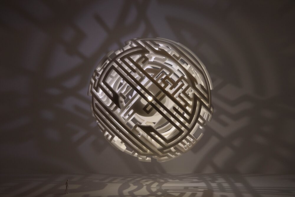 White model of intricate sphere, suspended in space, with straight and curved lines. Light glows from within the model creating shadows of lines in the background.