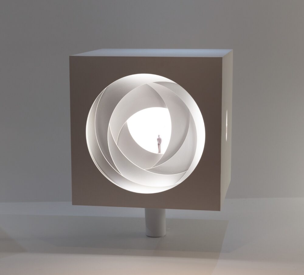 Cubic white model with a large circle in the center of one side made of layered cutouts. A tiny figure stands on one of the layers. White light glows from within the cube.