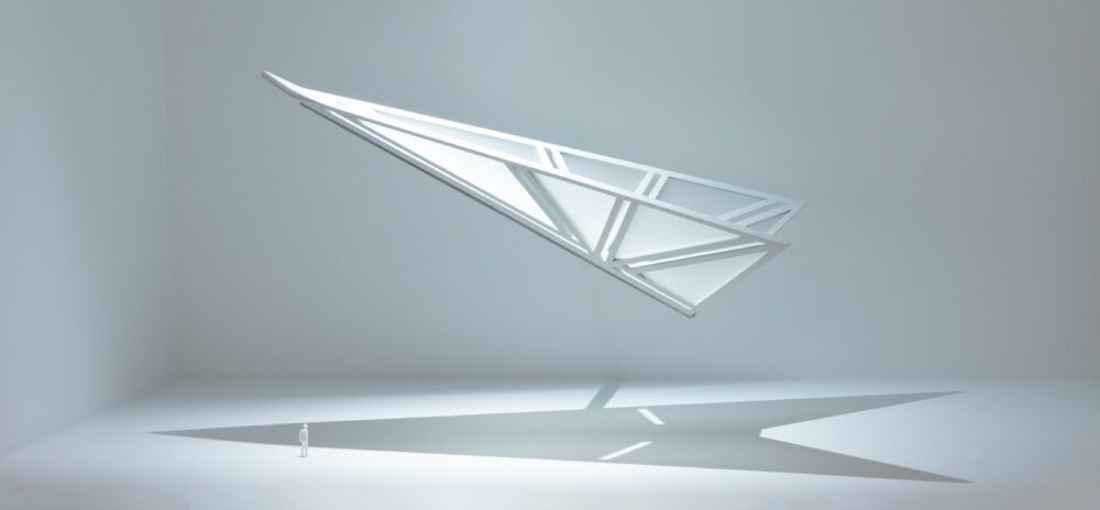 White model of a geometric form that resembles a paper airplane made of small segmented triangles that are translucent; the model is suspended in a white space and is spotlit.