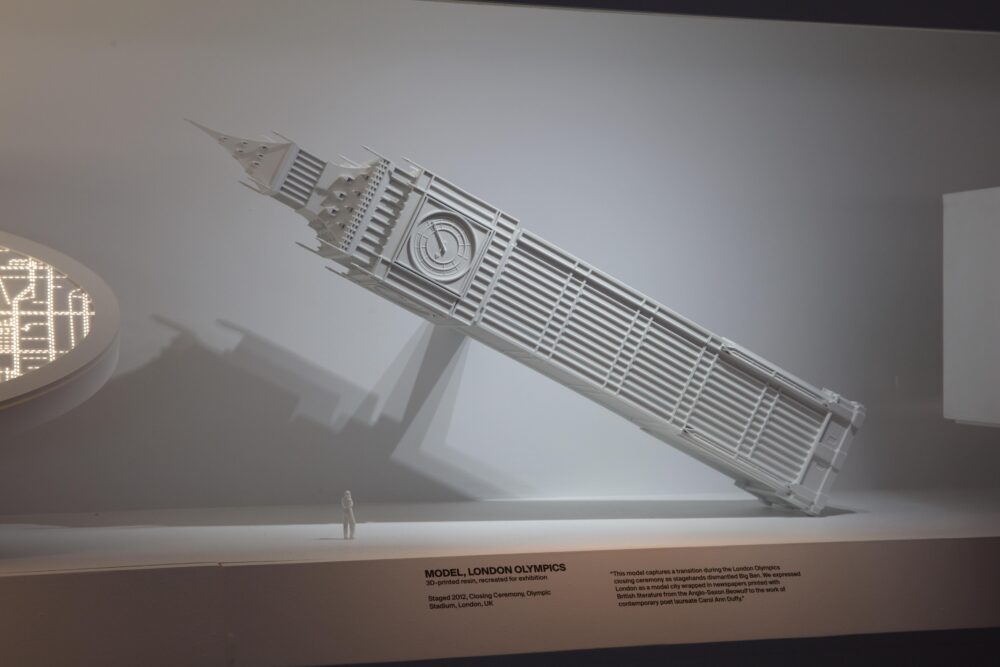 Installation view of a white model of a large clocktower, resembling Big Ben, that is tilted to the left at an extreme angle, almost laying on its side.