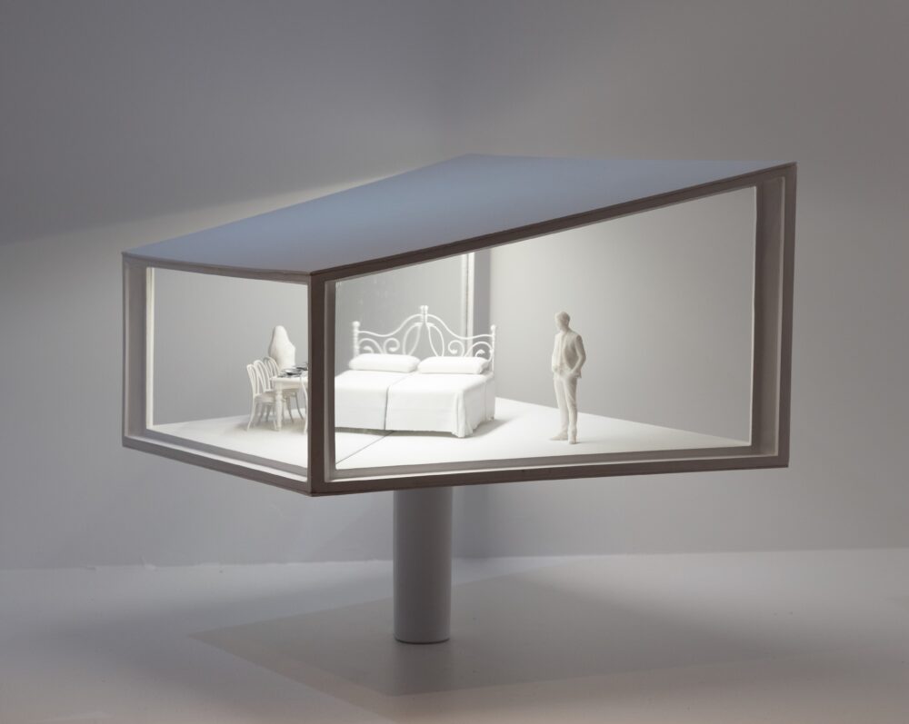 White model of a trapezoidal structure with white light glowing from inside, illuminating a bed, figure, and chairs.