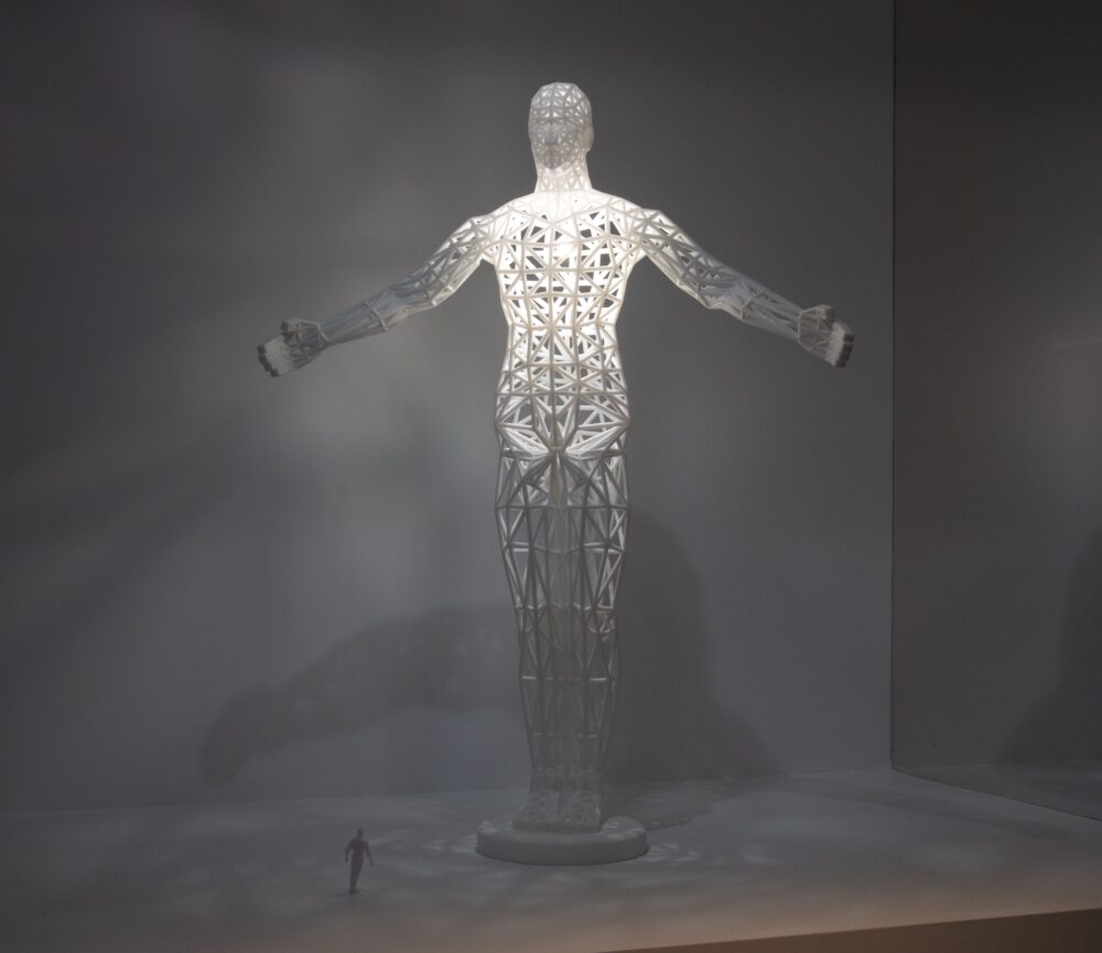 A large-scale white model of a standing figure with extended arms; white light glows from within the model revealing openings in the structure. A much smaller figure stands next to the model for scale.