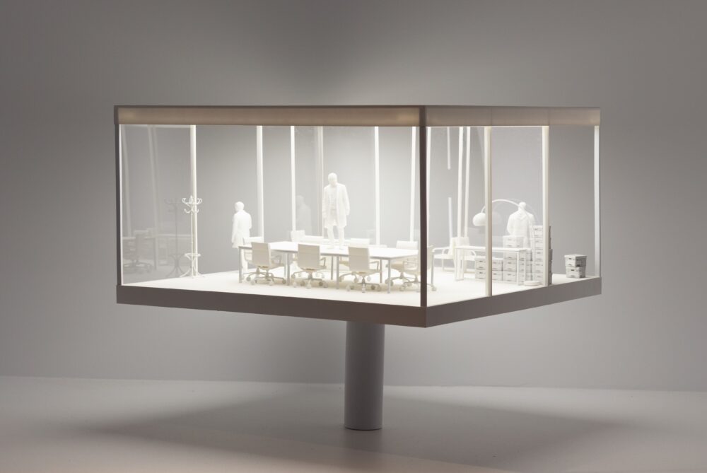 Elevated on a thin pole is a model of a midcentury-styled office with a table, chairs, and boxes. The space is inhabited by three small figures.