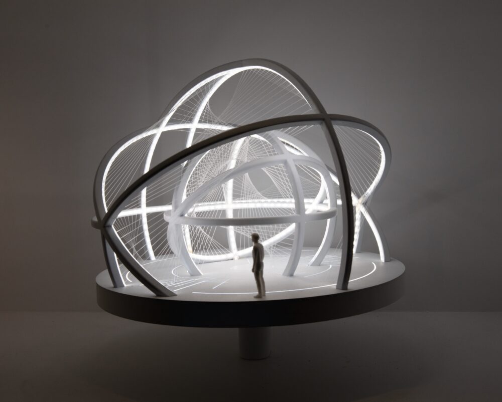 White model on a circular base of curved arches at different heights and angles forming two differently sized spherical structures, one inside the other. Delicate threads connect the two structures. Light glows from within the model; a figure stands at the edge of the model for scale.