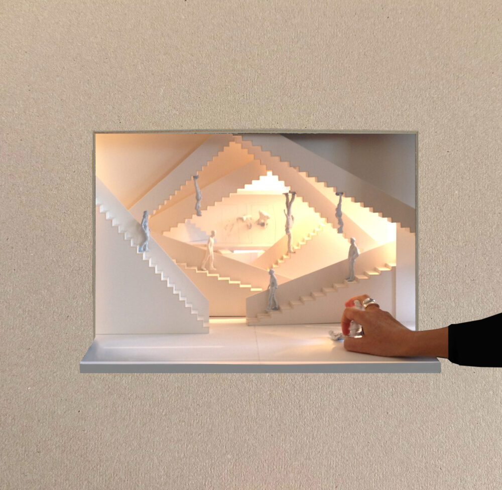 White model for a stage design with a person’s arm and hand in the foreground showing scale. The model is made of repeating layered staircases at different angles and small figures walking along them; a glowing light illuminates the model from within.