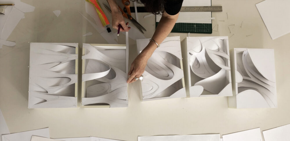 Top view of a person at a work table measuring and cutting paper to create curved forms within rectangular sculptures.