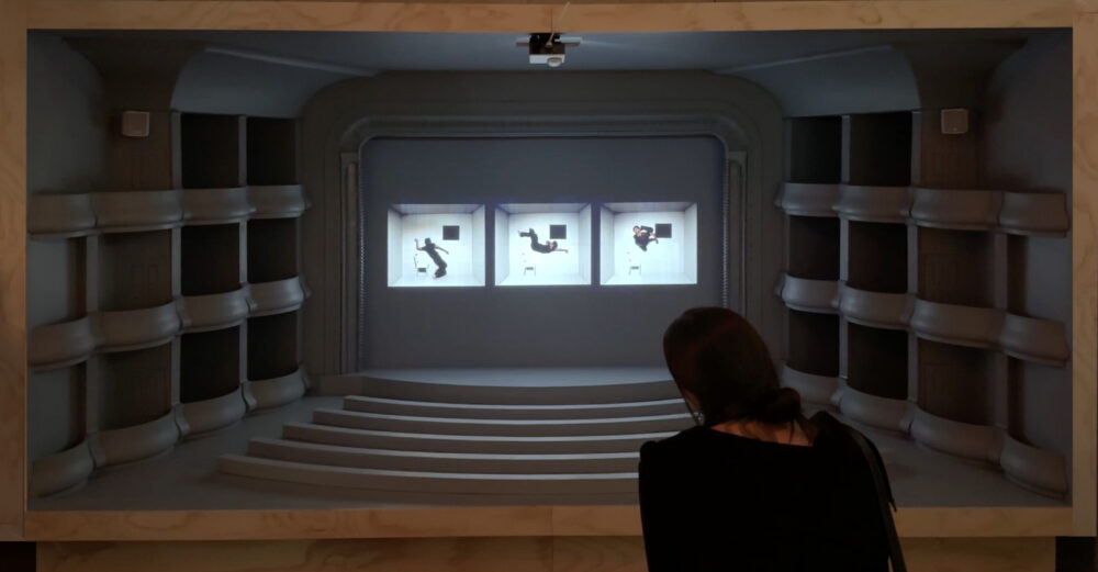 A figure is seen from behind looking into a large wooden crate in which a theater space is recreated with a projection on the theater screen of three white rooms lined up next to each other with a figure in each.