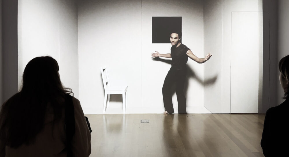 A figure is seen from behind looking at a long white wall with an image of a person in a white room next to a white chair being projected onto the wall.