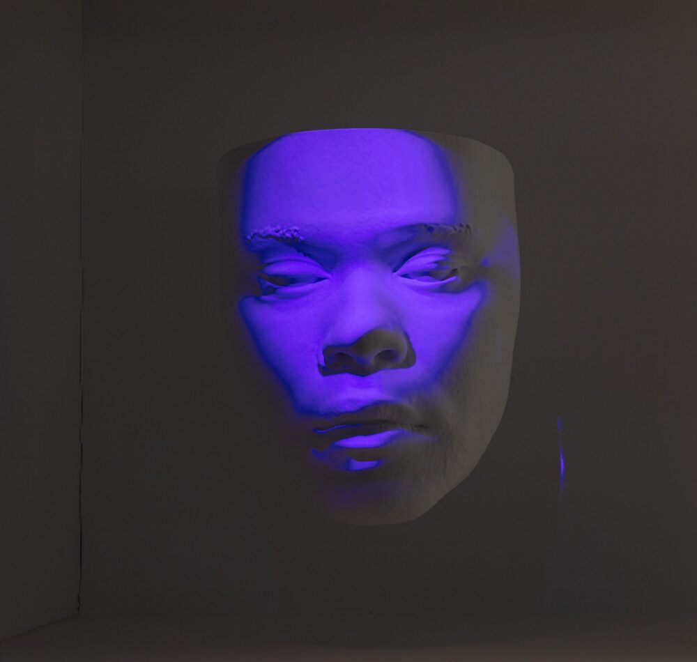 White model of a face suspended in air; purple light is projected onto the surface of the face.