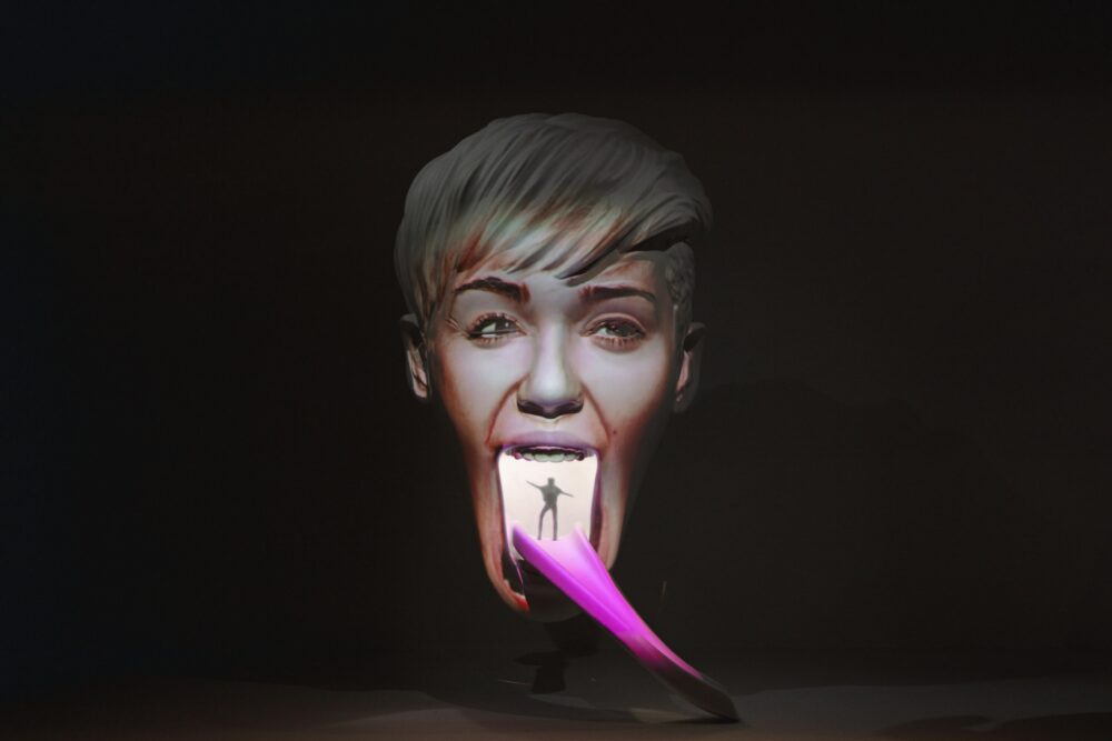 Model of a face with its mouth wide open and a long tongue extending downwards to the right. A colorful projection makes the hair blond and tongue pink. White light is glowing from inside the model revealing a small figure standing at the start of the tongue.