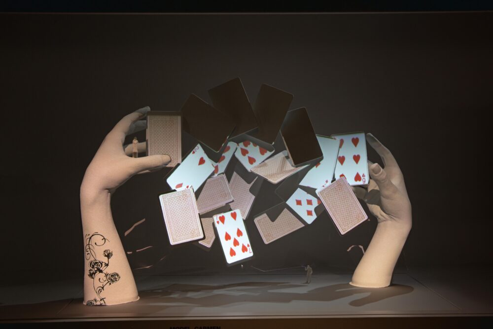 Model of two outstretched hands between which playing cards in red suits flutter.