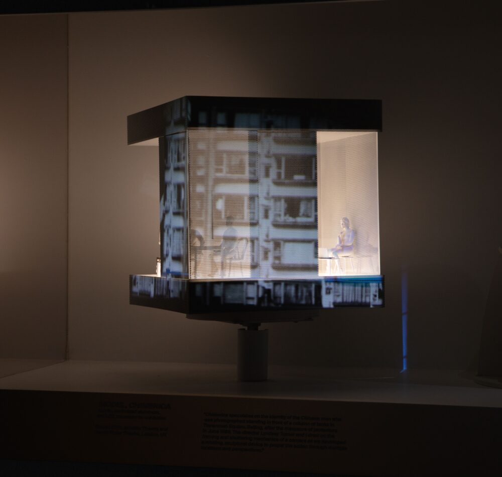 Installation view of a cubic model with an illuminated white interior space; projections of photos illuminate the exterior. A figure sits in a chair in the interior space.