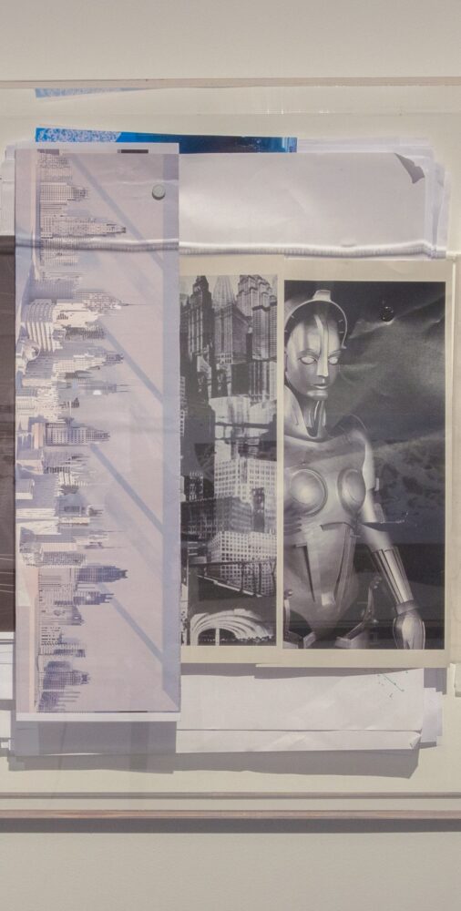 A pile of differently-sized papers pinned to a wall; one shows a city skyline and another shows a robotic figure.