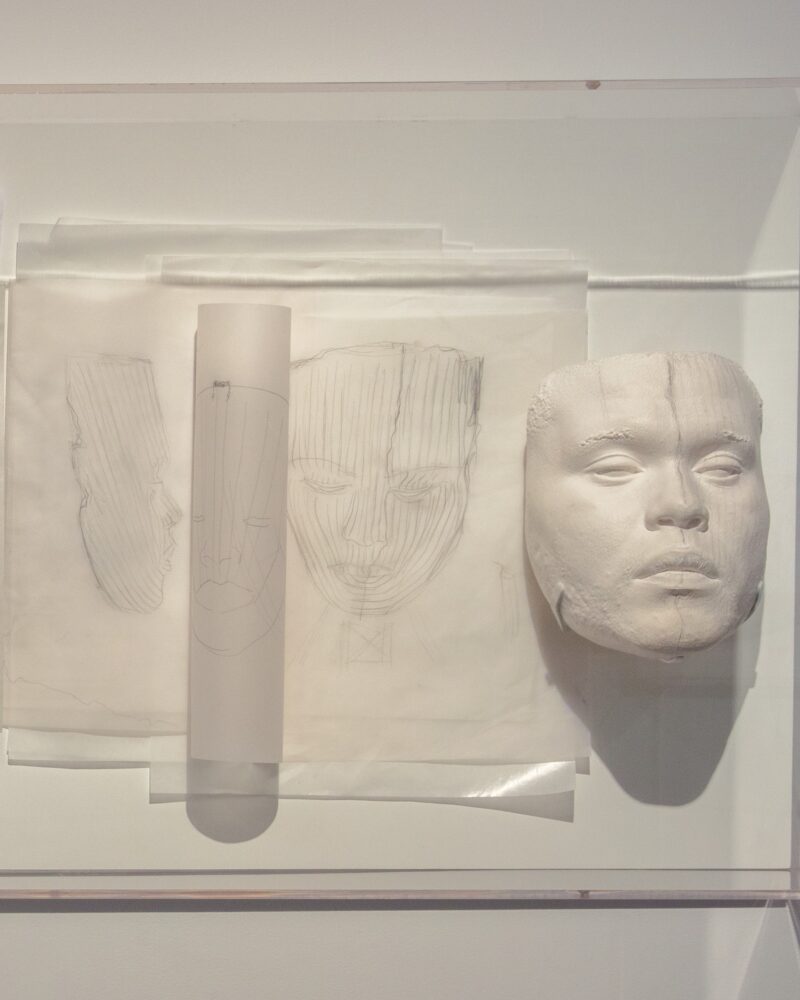 Several objects pinned to a wall: papers with drawings of a face, a translucent cylindrical object, and a sculpture of a face made in a white material.