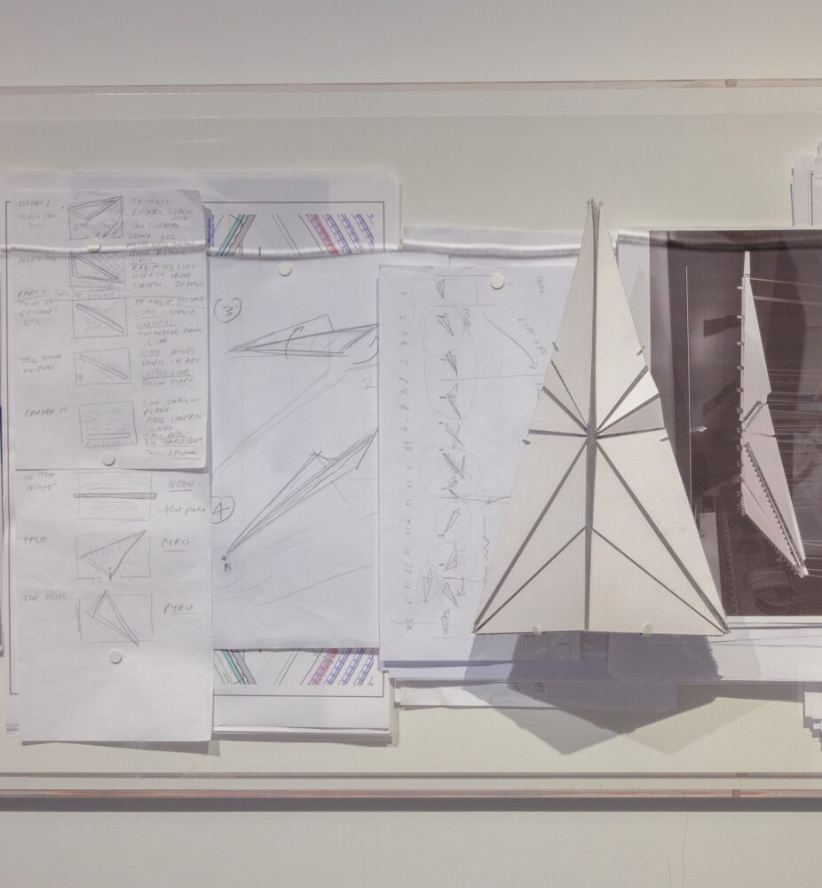 Several pieces of paper pinned to a wall depicting plans for a paper plane; a similar 3D triangular form is also pinned to the wall.