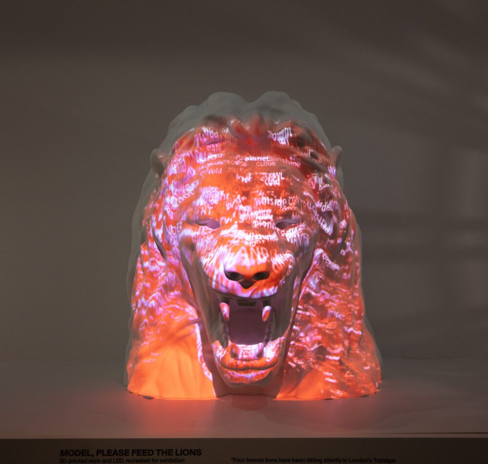 Straightforward view of a white model of a roaring lion’s head with red light and pink text projected on top.