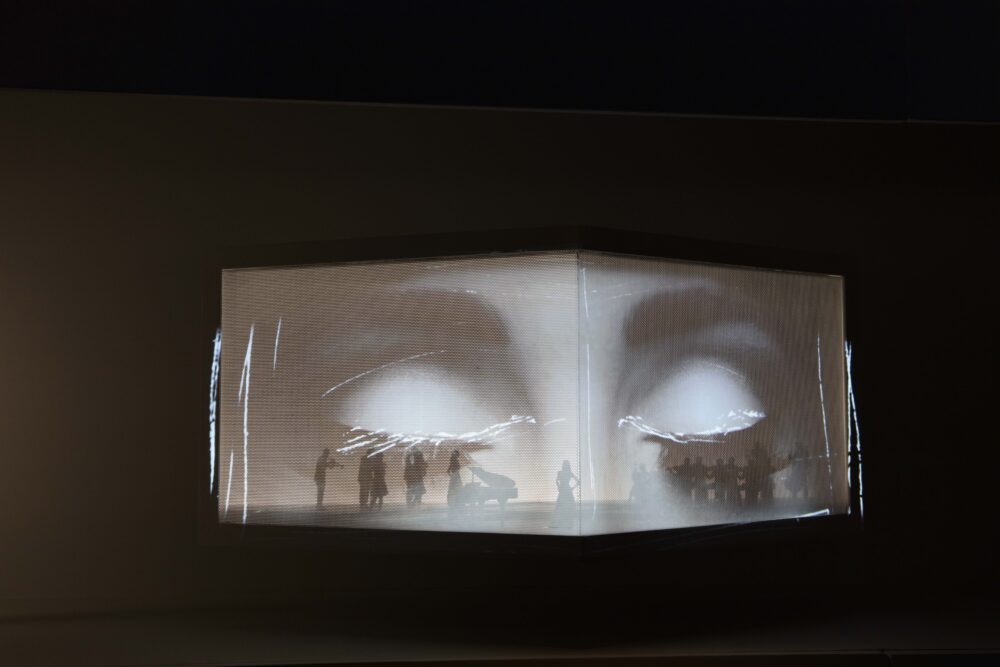 Slightly folded rectangular structure, above black background, with large-scale projection of two closed eyes; inside the structure are faint small figures.