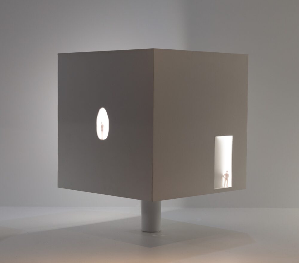 Cubic white model with white light glowing from within. Two sides are shown: the left one has a tiny figure standing on a small oval cutout in the center and the right has a tiny figure standing on a small rectangle cutout in the bottom right corner.