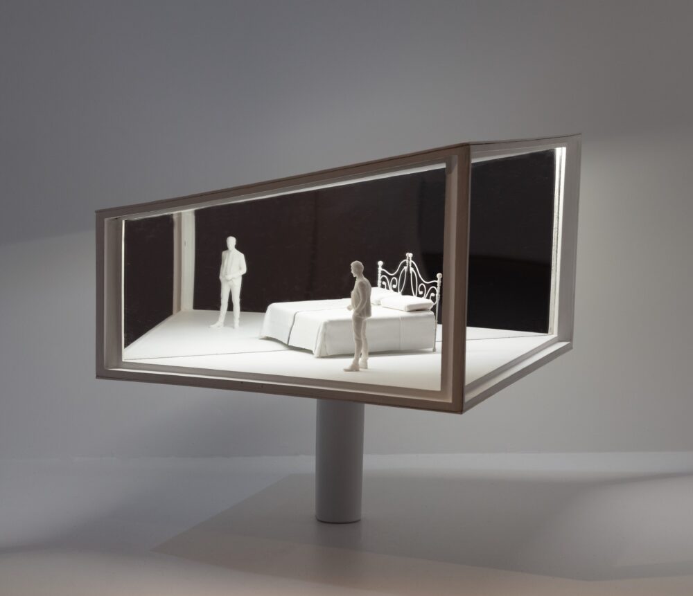 White model of a trapezoidal structure with a mirrored wall and white light glowing from inside, illuminating a bed and two figures standing on different sides of the space.
