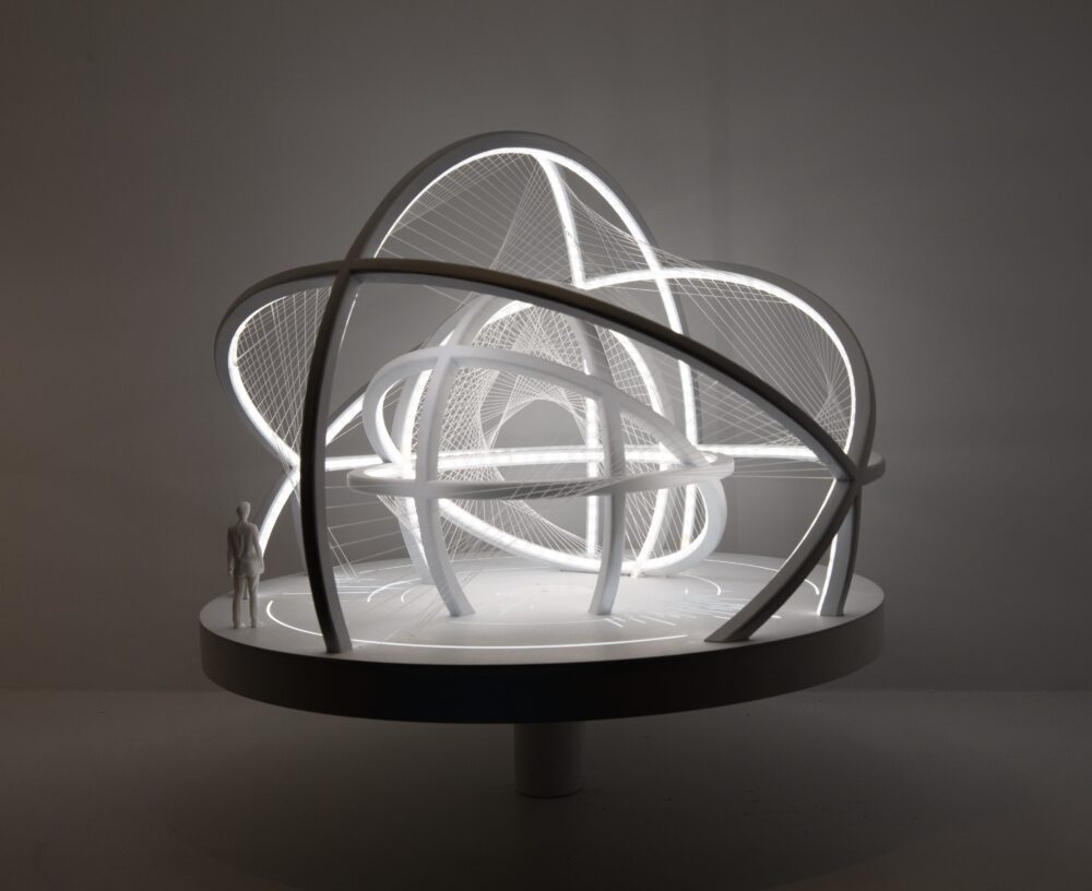 White model on a circular base of curved arches at different heights and angles forming two differently sized spherical structures, one inside the other. Delicate threads connect the two structures. Light glows from within the model; a figure stands at the edge of the model for scale.