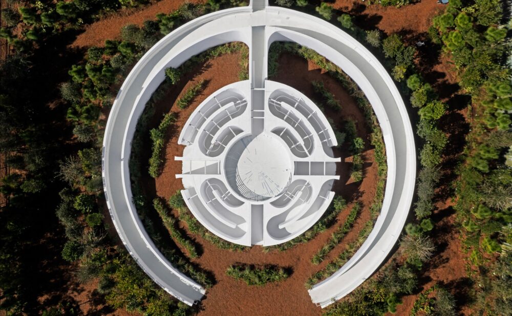 Aerial view of a white architectural structure situated outdoors and surrounded by trees; an open-ended circle structure is attached to a circle structure with winding paths in each quadrant; a round courtyard centers the structure.