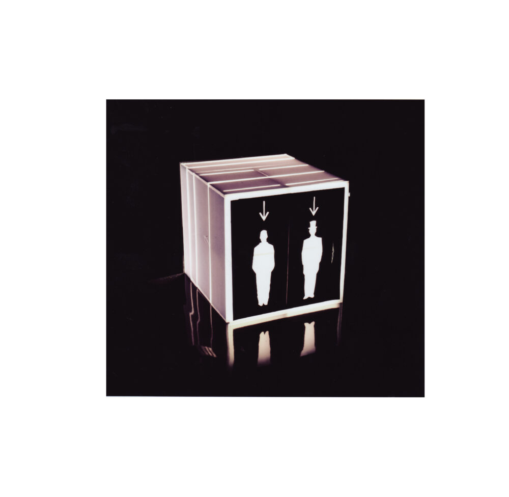 Three sides of a cube that emits light are shown before a black background; one side is black with silhouettes of two people standing.