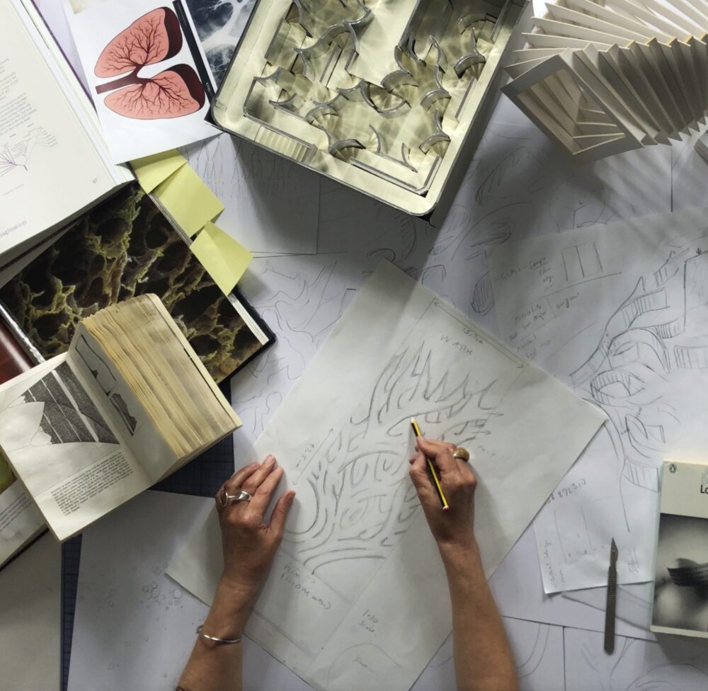 Top view of a person’s arms and hands drawing at a work table surrounded by drawings, books, and a geometric sculpture.