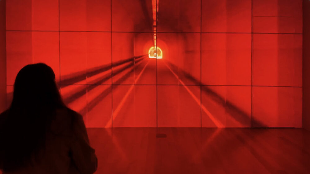 A figure is seen from behind looking at a long white wall divided into three rows of squares; projected onto the wall is a tunnel bathed in red light.