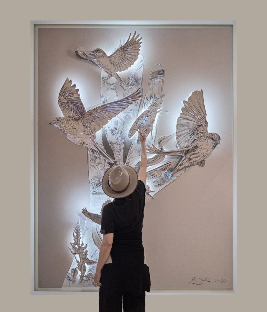 A person dressed in black stands in front of a large-scale artwork in a rectangular frame; the artwork is a paper sculpture of cutouts and black-and-white illustrations of realistically rendered birds forming the shape of a hand; white glowing light emphasizes the different layers and protruding cutouts.