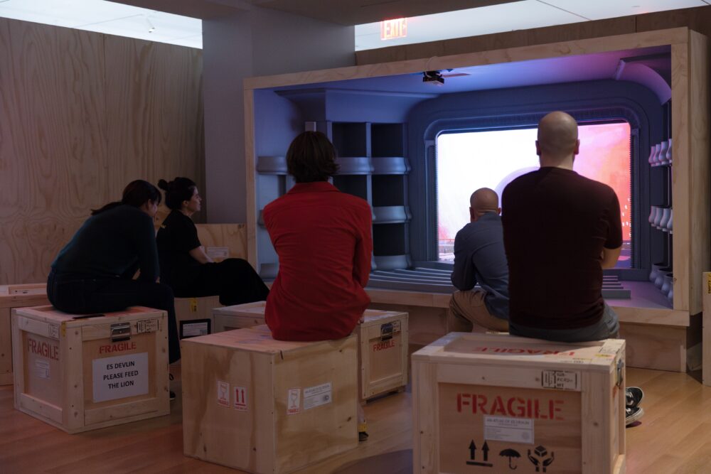 A group of people sit on different wooden crates facing a larger crate in which a theater space is recreated with projections of light on the theater screen.