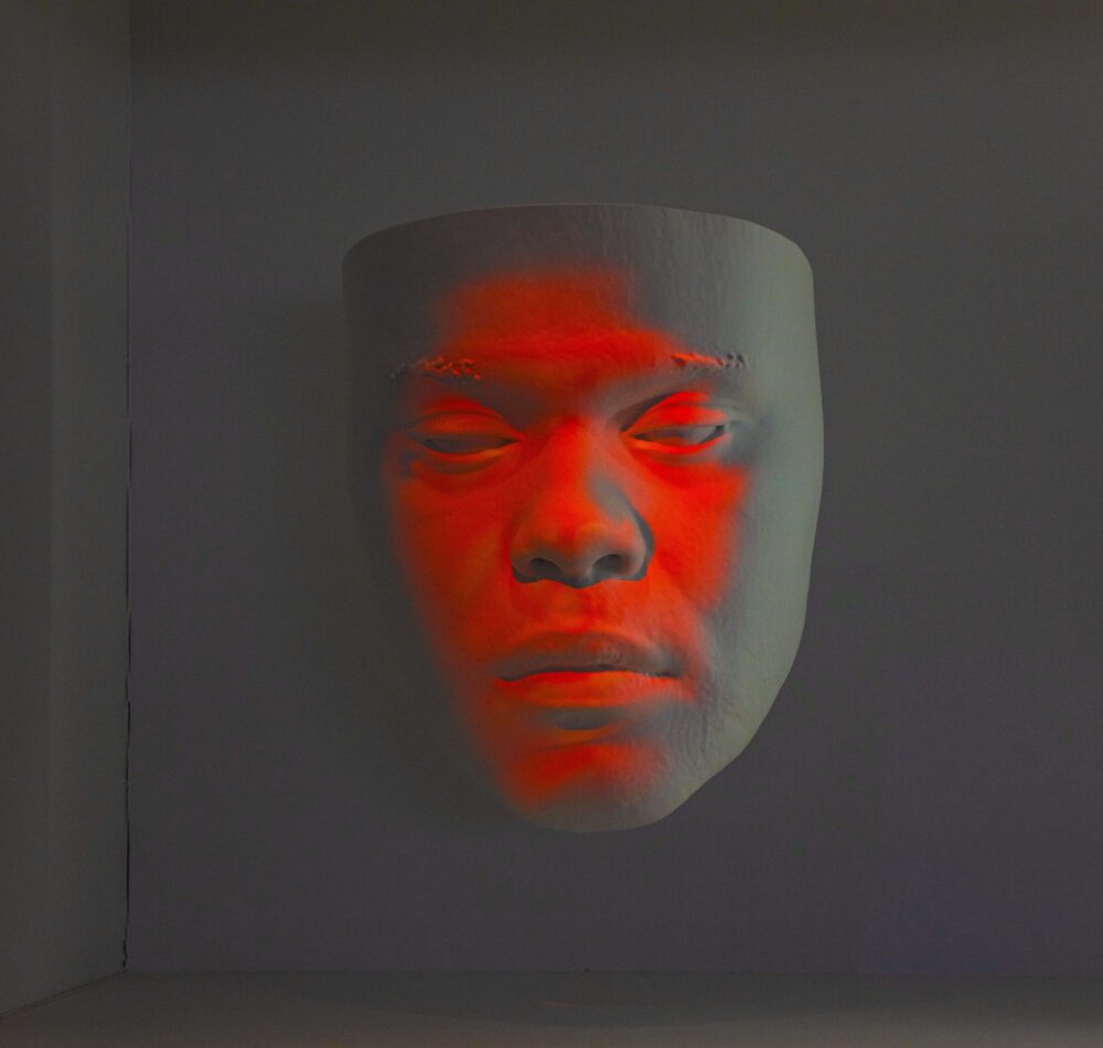 White model of a face suspended in air; red light is projected onto the surface of the face.
