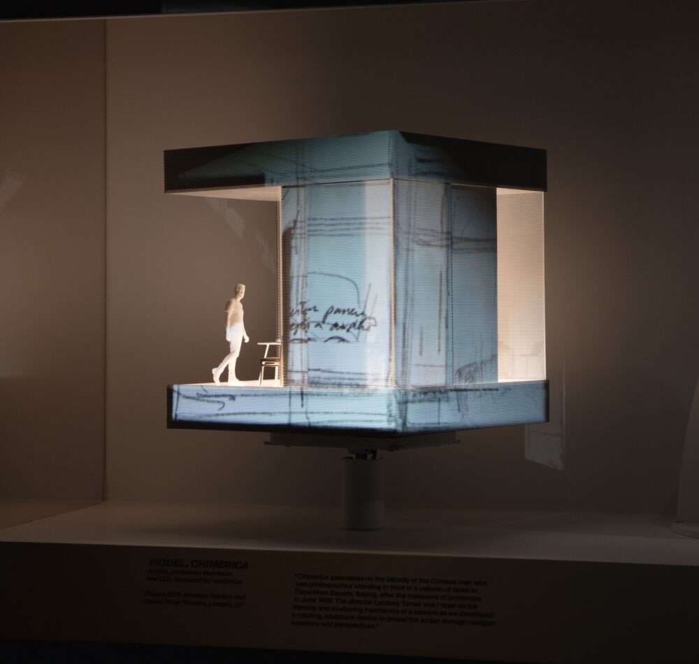 Installation view of a cubic model with an illuminated white interior space; projections of drawings illuminate the exterior. A figure stands in the interior space.