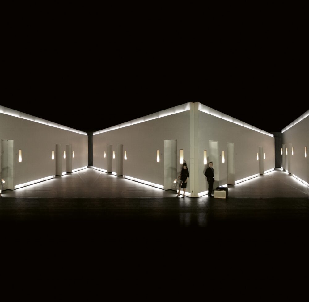 Panoramic view of two white triangular hallways in a darker space. White doorways and lights decorate the walls. Two people lean against the corner separating both hallways.